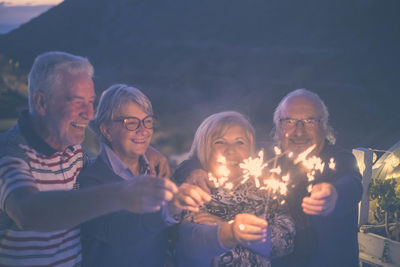 Cheerful senior friends holding lit sparklers at balcony during dusk