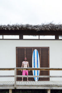 Young man standing by surfboard in front of door