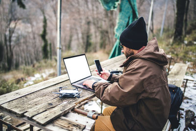 Side view of male hiker using mobile phone and laptop computer on table in forest
