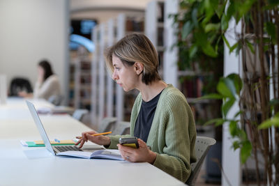 Focused female university teacher using laptop while sitting in quiet cozy library