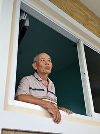 Low angle view of mature man looking through window while standing in house