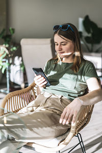 Young pretty woman in green beige clothes sitting in wicker chair in sunlight using smartphone