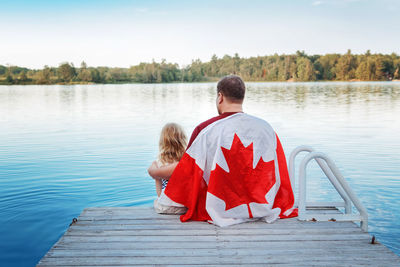 Father and daughter wrapped in large canadian flag sitting on wooden pier by lake. canada day 