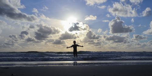 Silhouette boy standing with arms outstretched in sea against sky