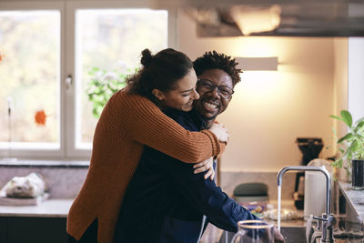 Side view of happy woman embracing man washing hands in kitchen at home