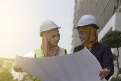 Happy female architects looking at each other while holding blueprint against clear sky
