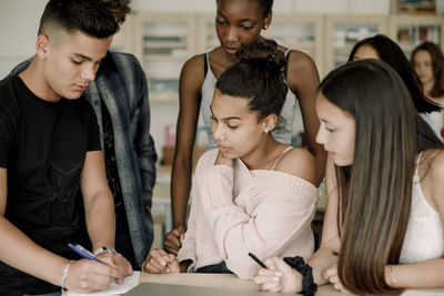 Teenage boy solving while friends looking in book by table in classroom