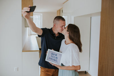 Smiling young couple kissing with digital tablet while photographing at home