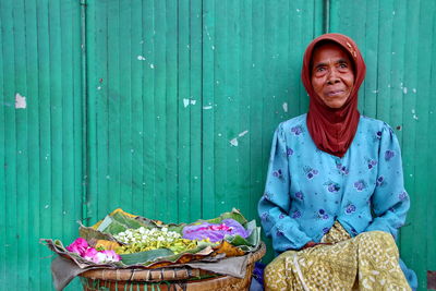 Smiling woman wearing headscarf selling flowers while sitting against wall