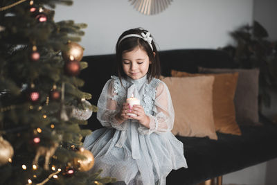 Cute girl holding candle sitting on sofa by illuminated christmas tree