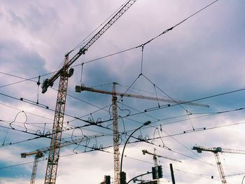 Low angle view of cranes and cables against cloudy sky