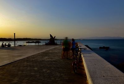 People riding bicycle on sea against sky during sunset