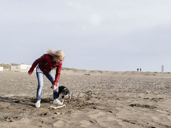 Full length of woman playing with dog on sand at beach against sky