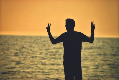 Silhouette man gesturing peace sign at beach against clear orange sky