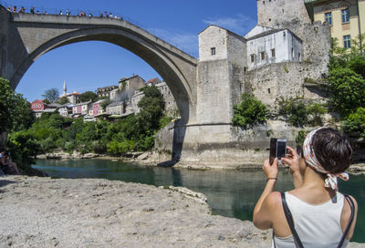 Rear view of woman photographing with smart phone by river in city during sunny day