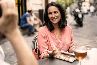 Smiling young woman talking to friend while sitting at sidewalk cafe