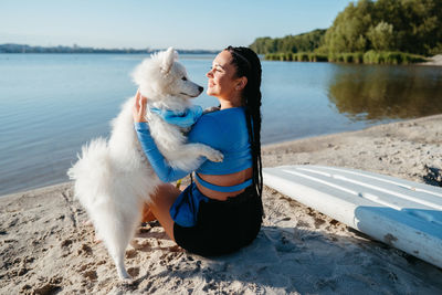 Woman with dreads hugging her best friend, snowy dog breed japanese spitz, on beach of city lake
