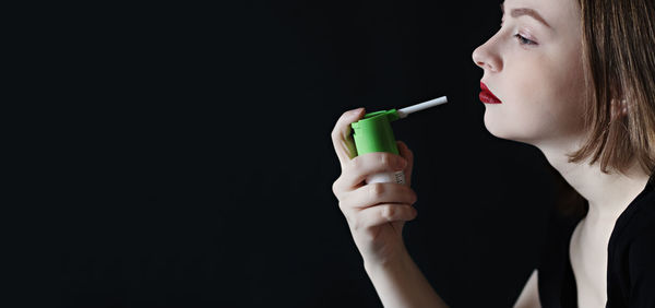 Close-up of young woman holding inhaler over black background