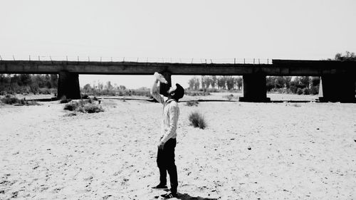 Full length of man drinking water from bottle against bridge on sandy field