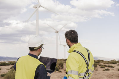 Engineers wearing reflective clothing working on laptop at wind farm