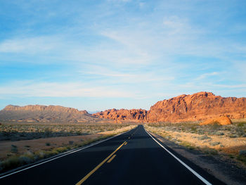 Country road amidst field leading towards rocky mountains against sky