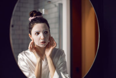 Woman in the bathroom in front of the mirror puts on a rejuvenating face mask from wrinkles
