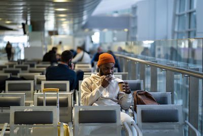 Young african man drinking coffee and eating sandwich while waiting for flying at airport terminal