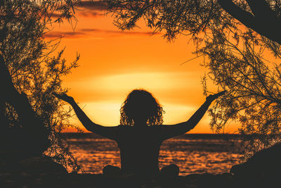 Rear view of silhouette woman with arms outstretched sitting by sea against sky