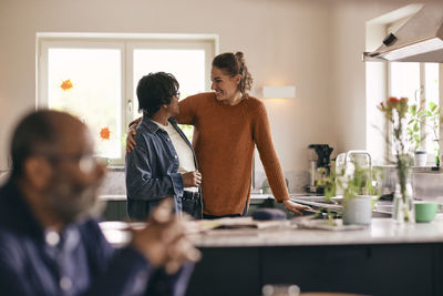 Happy daughter-in-law with arm around senior woman standing in kitchen at home