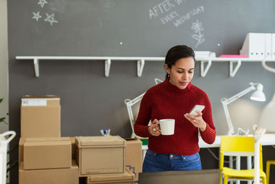 Businesswoman holding coffee cup while using smart phone against wall at creative office
