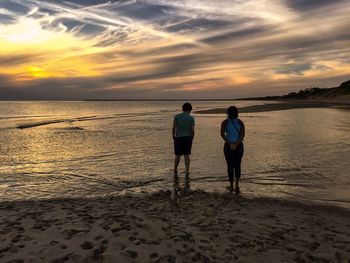Rear view of friends standing on beach against sky during sunset