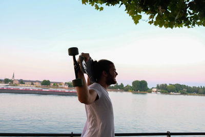 Side view of man holding skateboard while standing by lake against sky