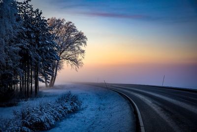 Road by snow covered landscape against sky during sunset