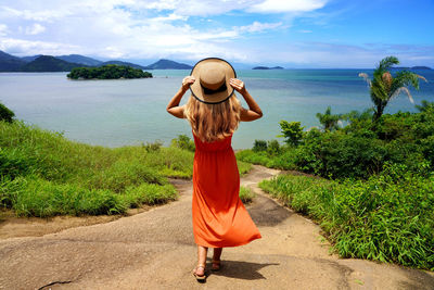 Beautiful girl with orange summer dress holding hat enjoying view of costa verde, brazil