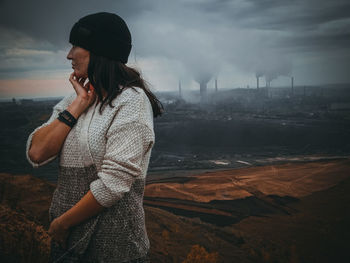Thoughtful woman looking away while standing against factory emitting smoke