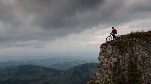 Man with bicycle on mountain against cloudy sky