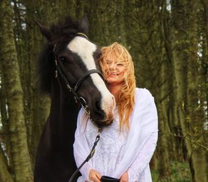 Smiling woman standing by horse against trees