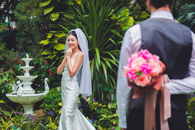 Groom hiding flowers while bride waiting against plants