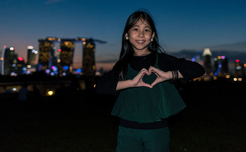 Portrait of girl making heart shape while standing in city at night