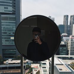 Reflection of man photographing with mobile phone on circular mirror against buildings