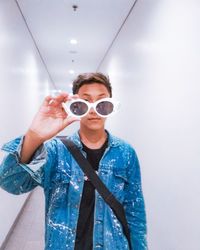 Young man holding sunglasses while standing amidst white wall