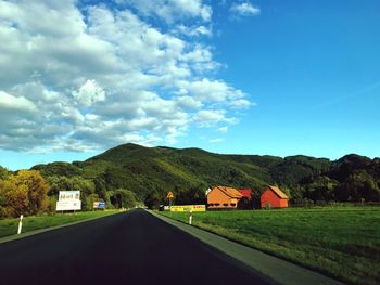 Road amidst field and houses against sky