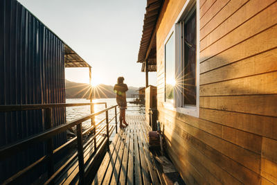 Man standing on railing by sea against sky during sunset