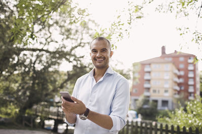 Portrait of happy businessman holding smart phone while standing at park in city