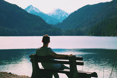 Rear view of man sitting by lake against mountains