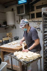 Side view of female baker in mask and shield standing at table in kitchen and making round shaped bread from raw dough while working in bakehouse