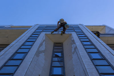 Low angle view of window washers working on building against clear blue sky