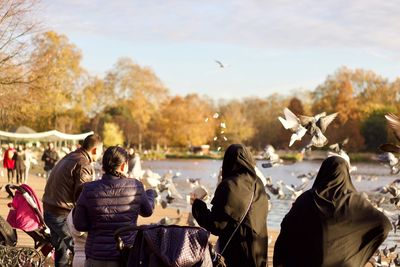 Rear view of people feeding birds by lake against sky