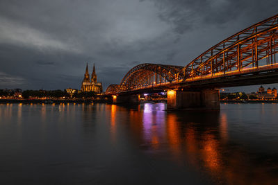 Illuminated bridge over river against cloudy sky