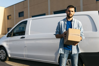 Young delivery man with package standing in front of van